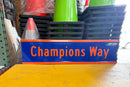 Blue sign with 'Champions Way' text on a yellow surface, surrounded by traffic cones and black plastic crates.